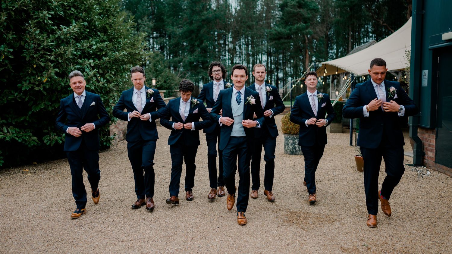 Groomsmen in matching navy suits walking confidently together outside a rustic wedding venue.