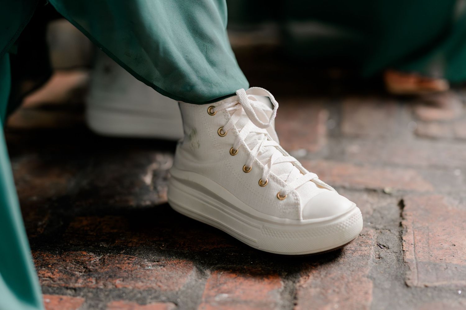 White high-top Converse sneakers with gold eyelets worn with green pants on a brick surface.