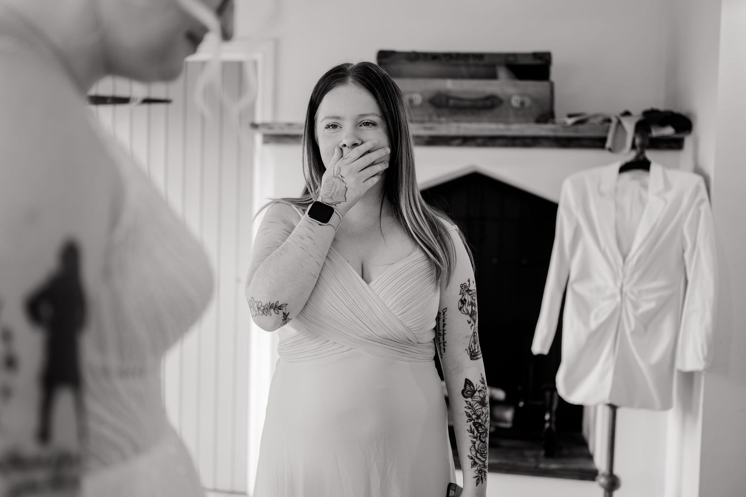 Black and white photo of a bride in a white dress covering her mouth in surprise while getting ready.