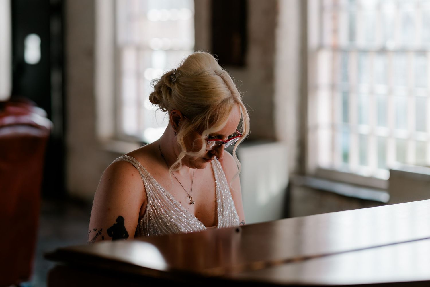 Blonde woman in elegant beaded dress playing piano by a window in soft natural light.