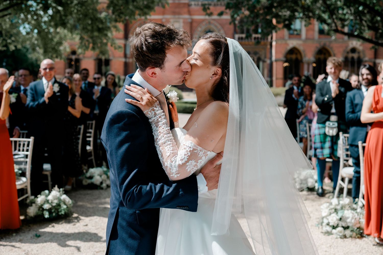 Newlywed couple sharing first kiss at outdoor wedding ceremony with guests applauding in background.