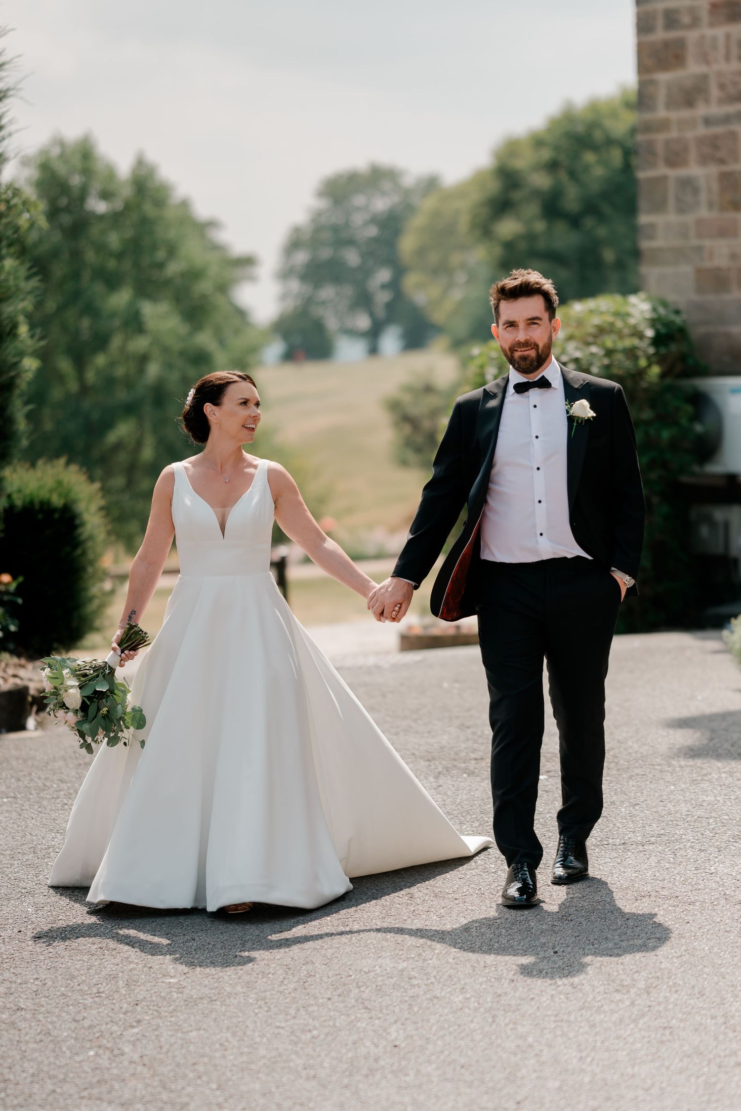 Bride in white gown and groom in black tuxedo walk hand in hand outdoors on their wedding day.