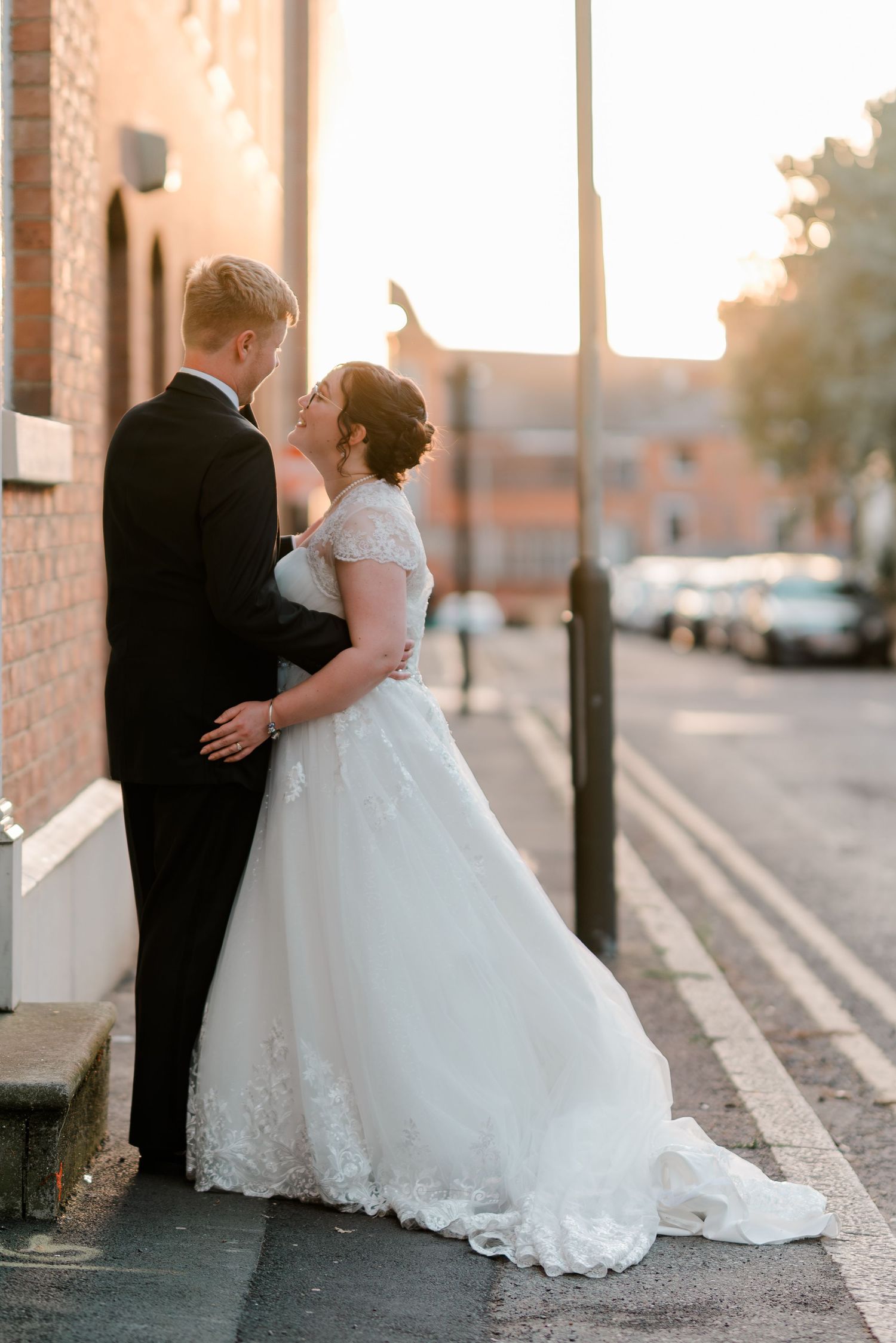 A bride and groom share a romantic kiss on a sunlit street, the bride wearing a lace ball gown wedding dress.