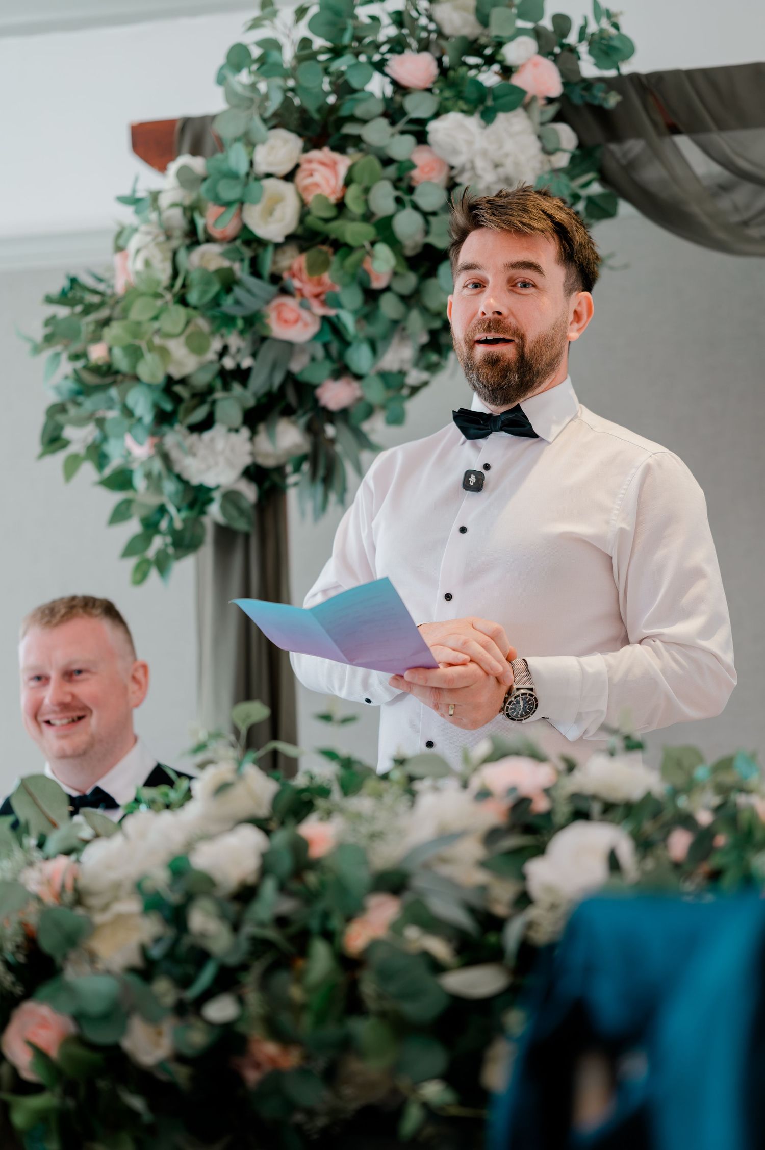 Bearded groom in white shirt and bow tie reads a speech from a blue notebook beside lush pink and white wedding flower arrangements.