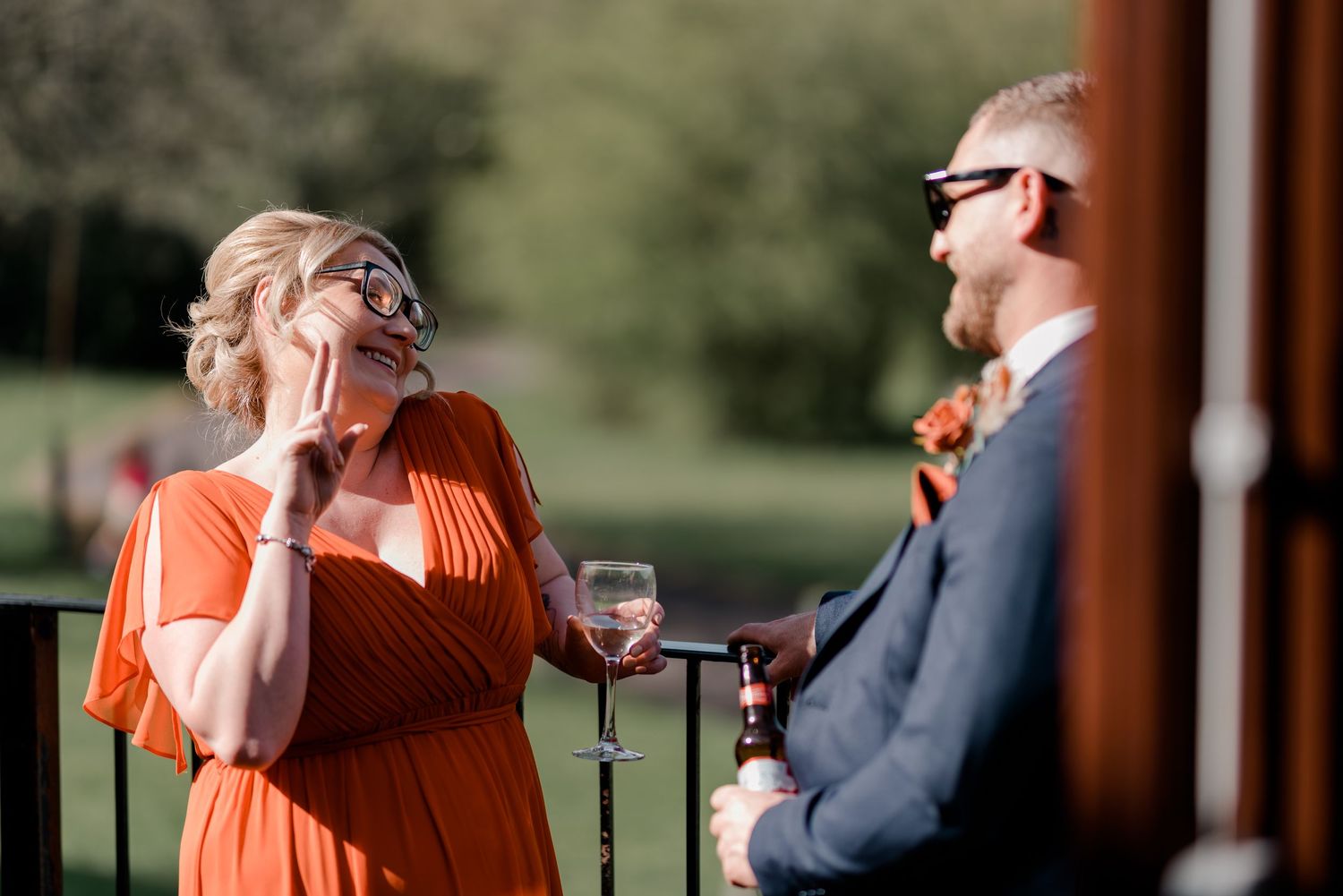 Two wedding guests in sunglasses share a laugh while holding wine glasses outdoors on a sunny day.