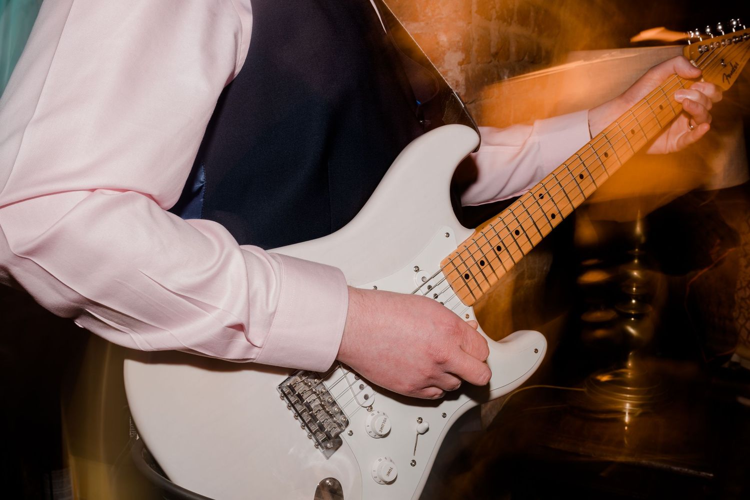 A musician plays a white Stratocaster electric guitar with a blurred background.
