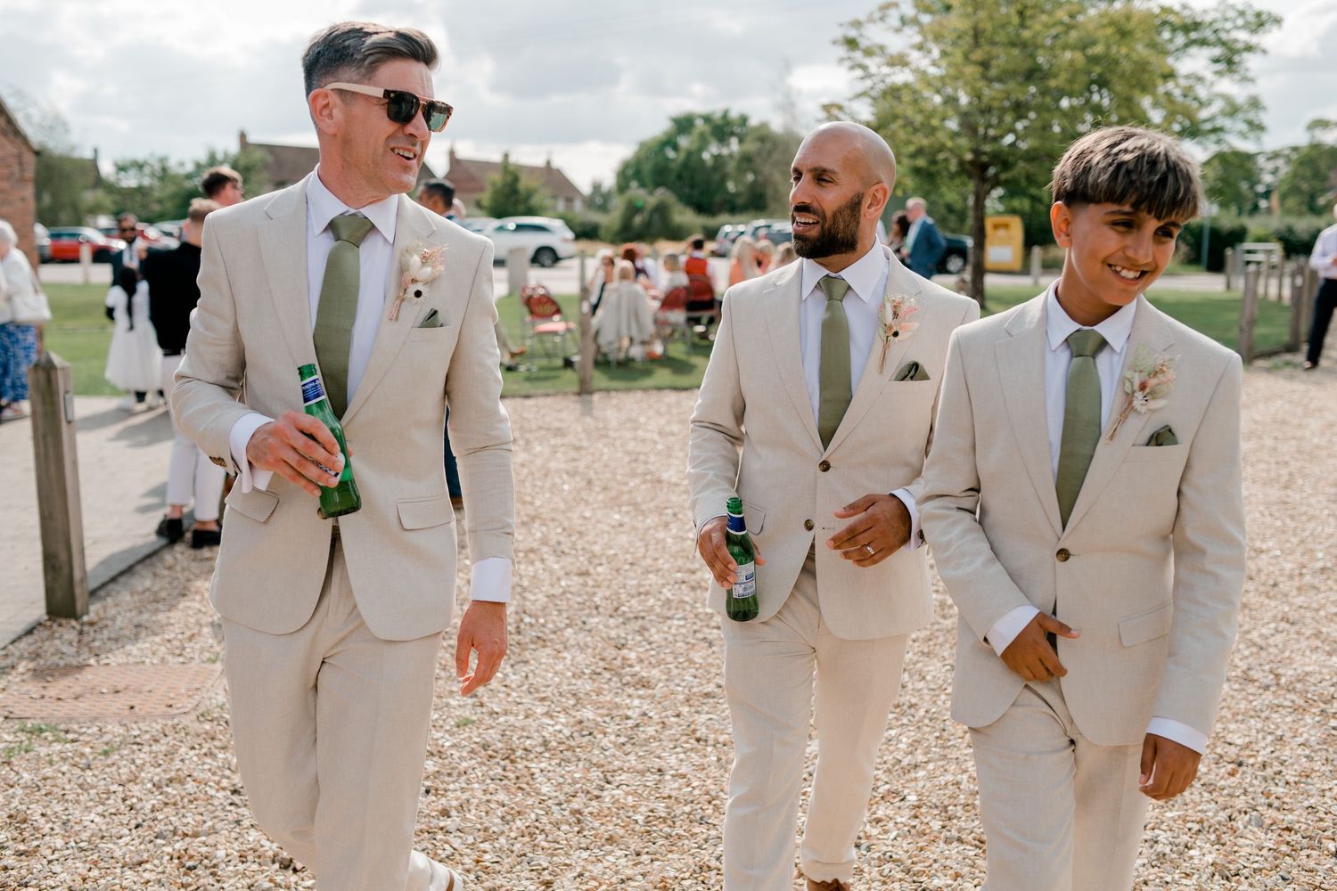 Groomsmen in beige suits walking and laughing while holding drinks at an outdoor wedding.