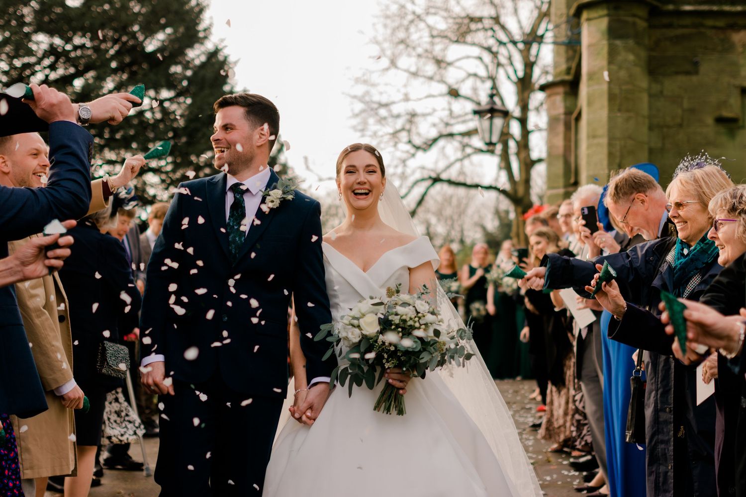 Joyful bride and groom walk through confetti shower outside historic church surrounded by cheering wedding guests.
