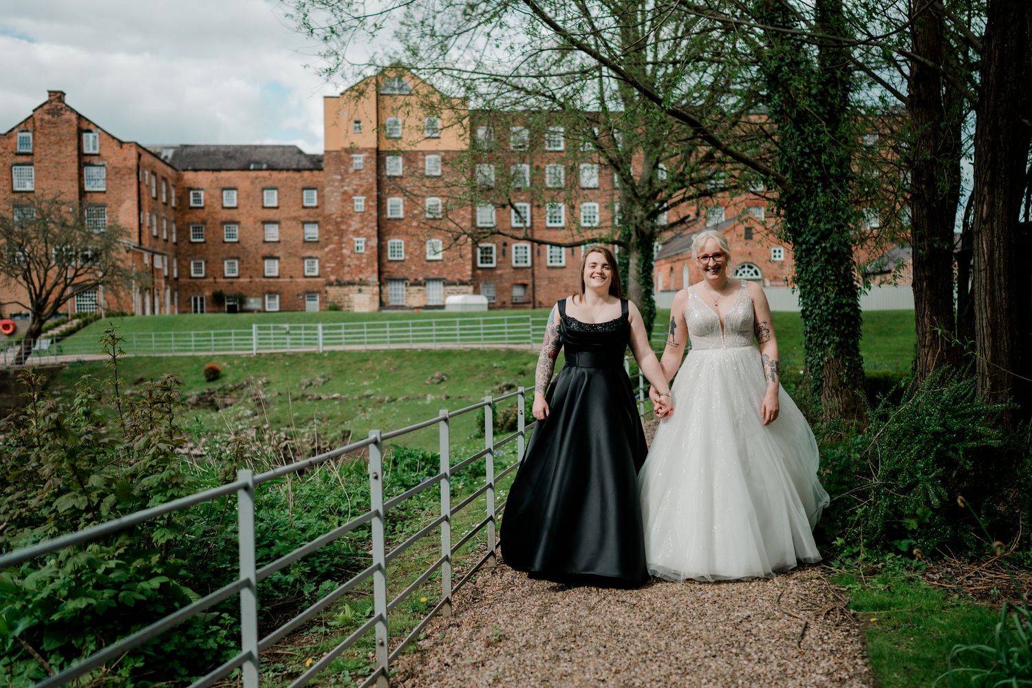 Two brides holding hands, one in black gown, one in white, walking path by historic brick mill building.