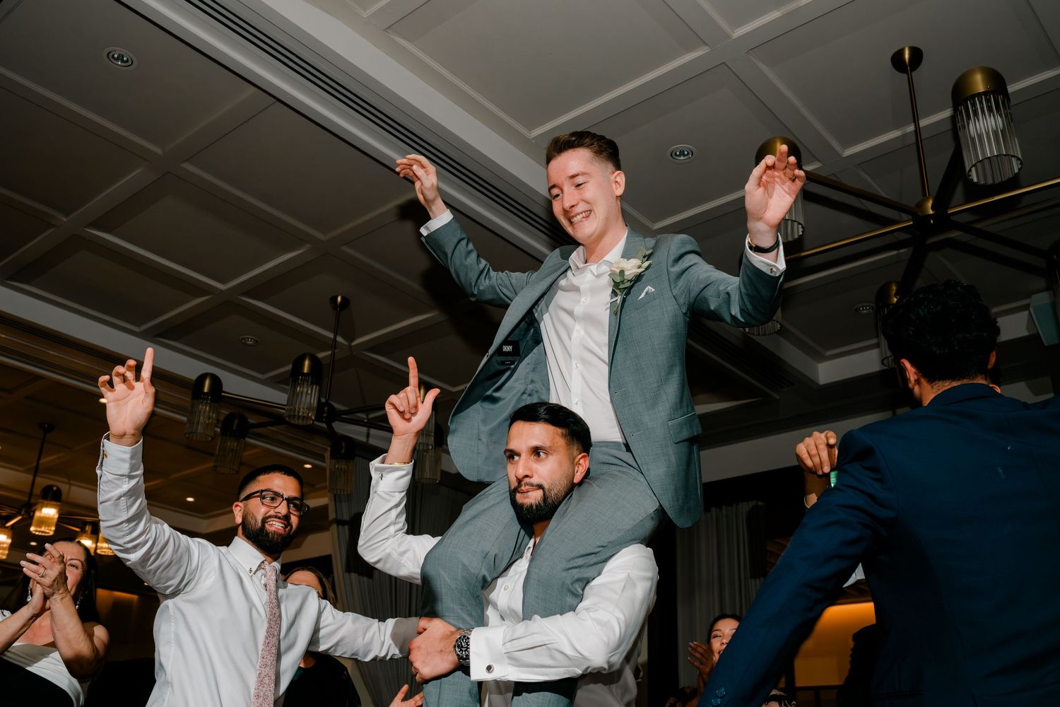 Groom in teal suit lifted on shoulders by groomsmen celebrating at wedding reception party.