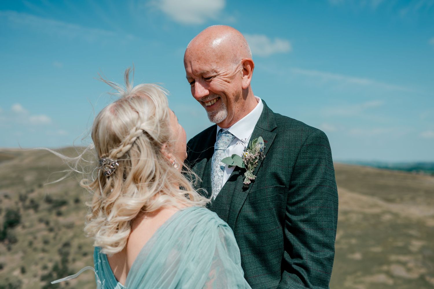 Smiling bald groom in dark suit with boutonniere gazes at blonde bride in teal dress outdoors.