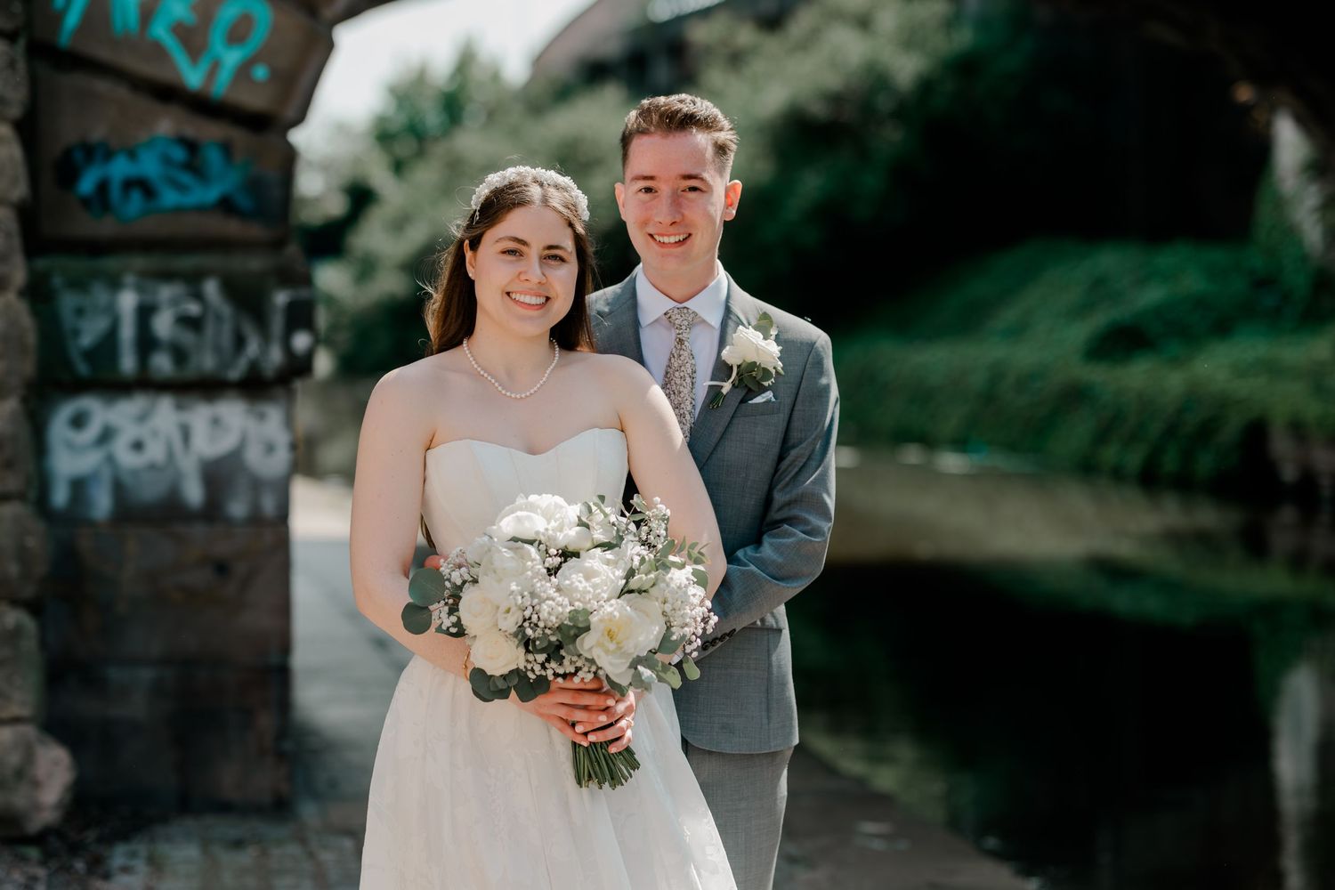 Bride and groom smiling together outdoors near a river, bride holding white floral bouquet in wedding attire.
