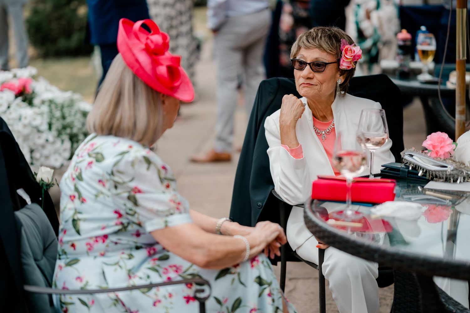 Two women in floral dresses and bright pink fascinators chat at an outdoor wedding reception table with flowers and champagne glasses.