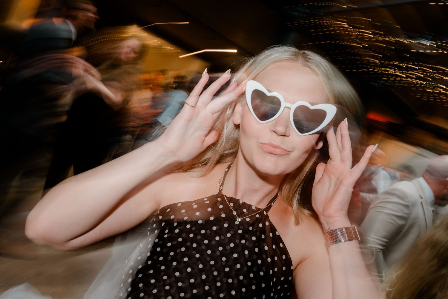 Blonde woman wearing heart-shaped sunglasses poses playfully on blurred wedding reception dance floor.