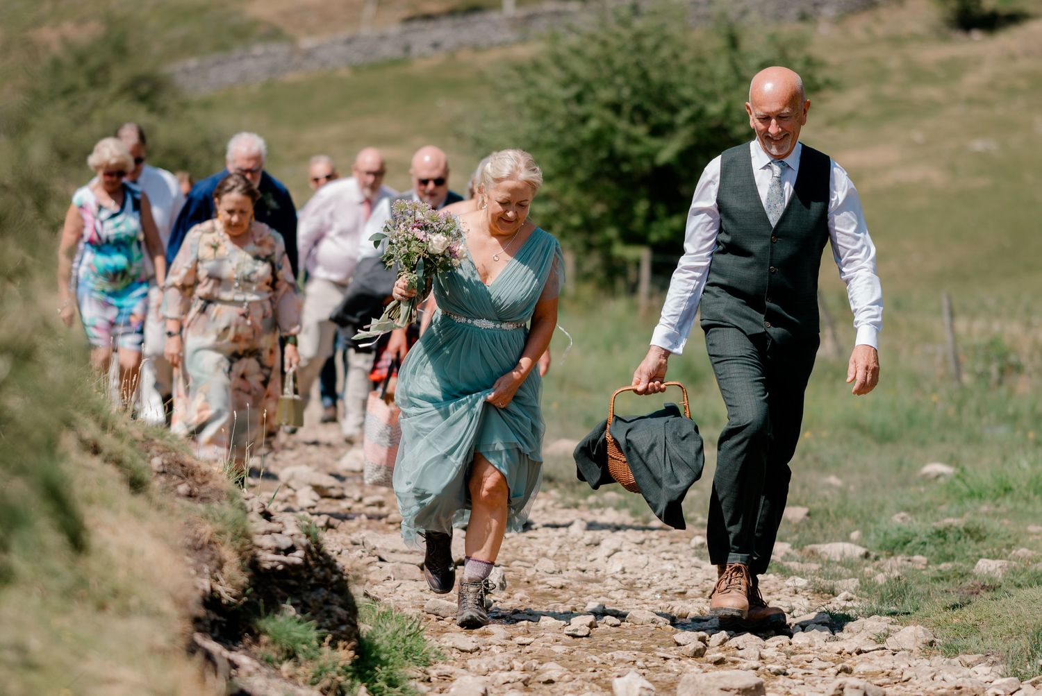 Wedding guests walking along a rocky path in sunny countryside, led by a couple in formal attire.