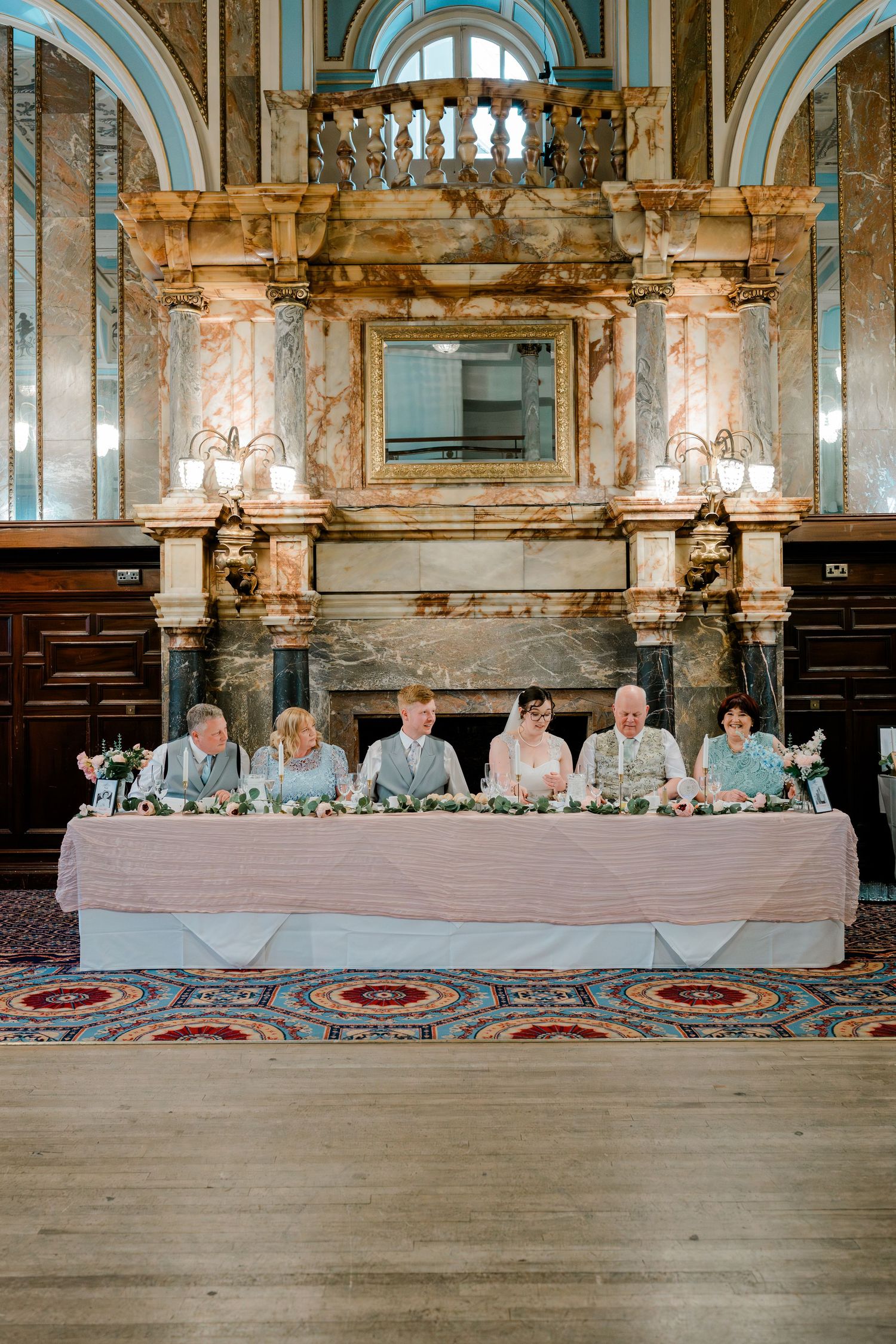 Wedding party seated at elegant head table with marble fireplace backdrop in ornate reception venue.