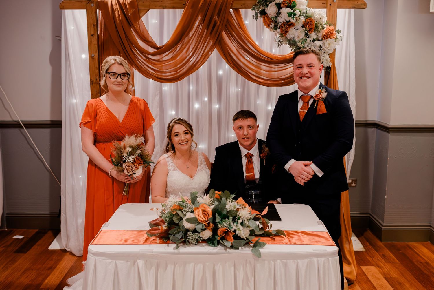 Wedding party stands behind signing table with orange decor and floral arrangements in banquet hall.