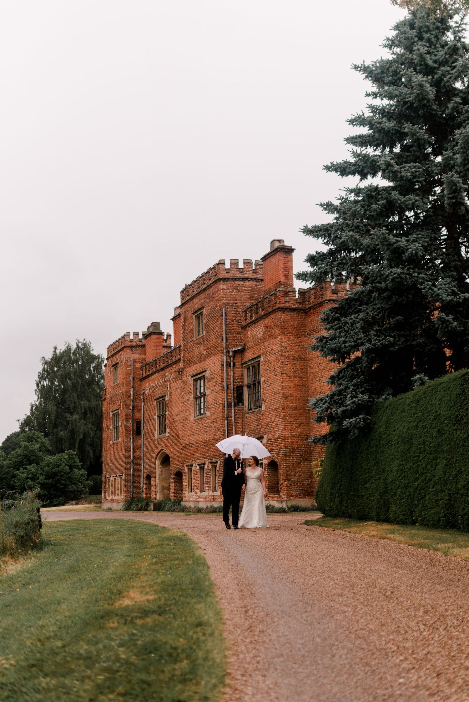 Historic red brick castle venue with gravel path and evergreen landscaping on overcast day.