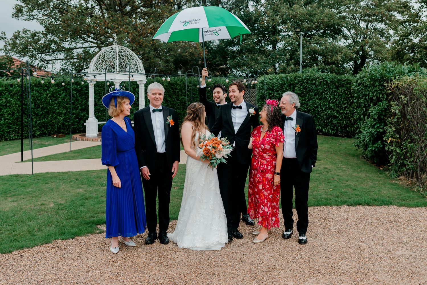 Wedding party members walking outdoors on a garden path under umbrellas on an overcast day.