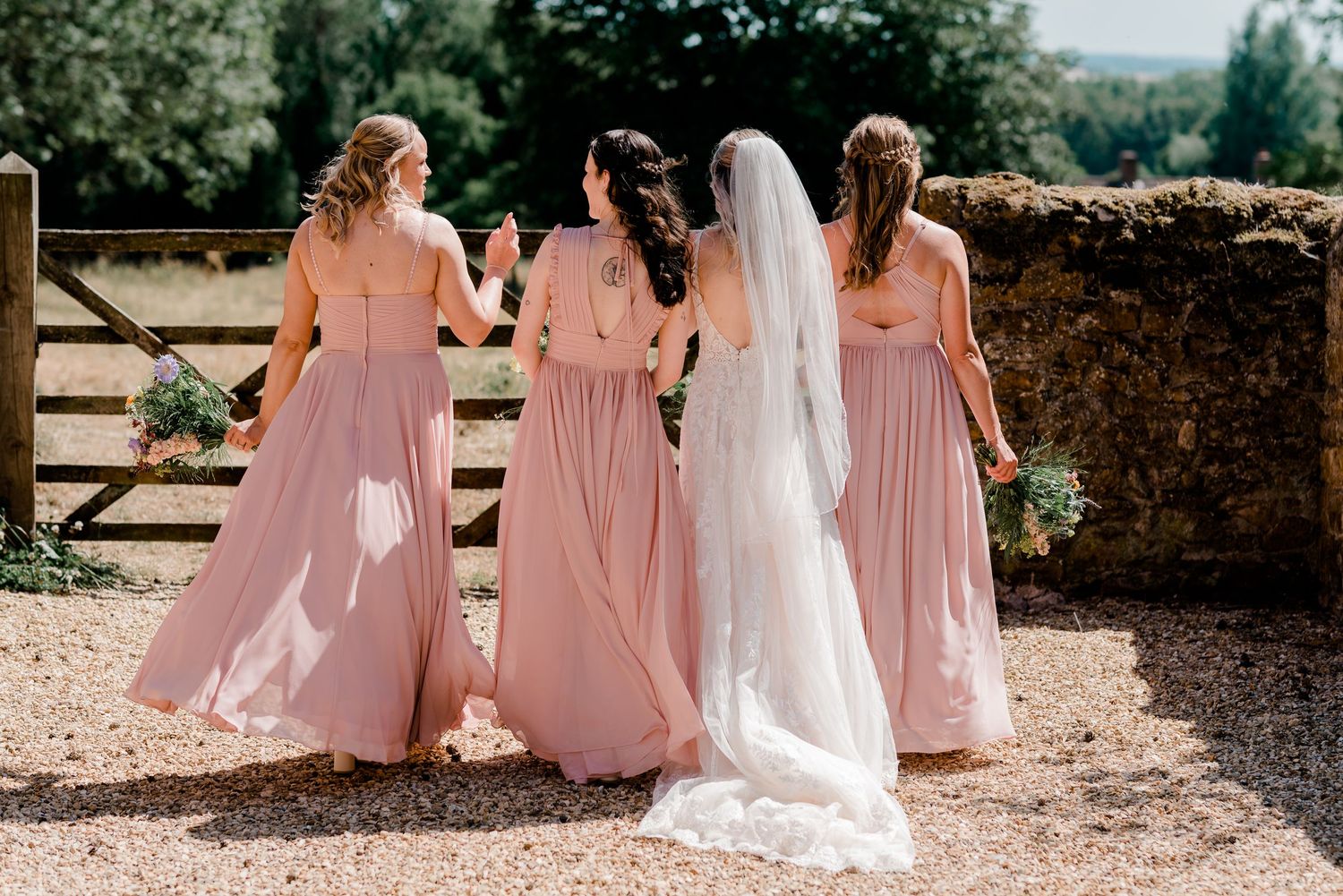 A romantic outdoor wedding scene with bridesmaids in pink dresses walking alongside a bride in white down a sunlit path.