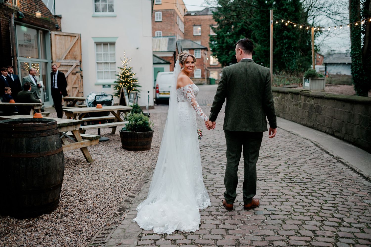Bride and groom walking hand in hand on cobblestone courtyard with festoon lights.