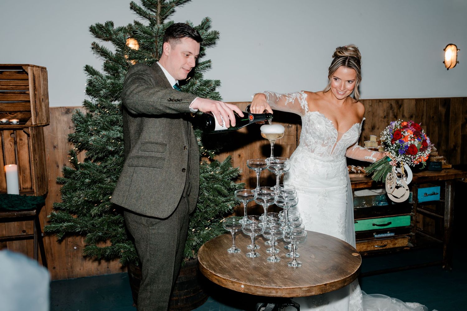 Bride and groom pouring champagne tower at winter wedding reception with Christmas tree backdrop.