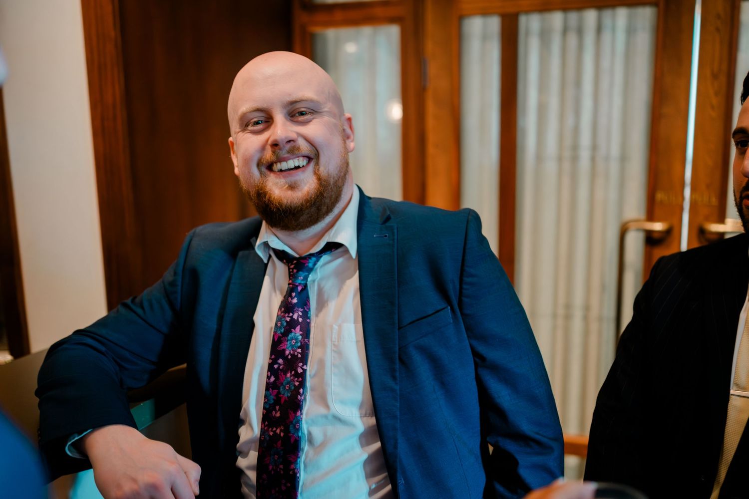 Smiling bald man in navy suit with floral tie at wedding reception.