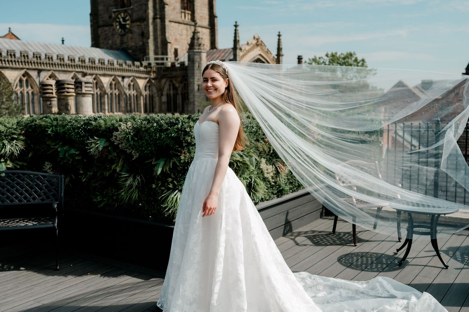 Bride in a lace ballgown with flowing veil caught in the breeze on a rooftop terrace beside a historic church.