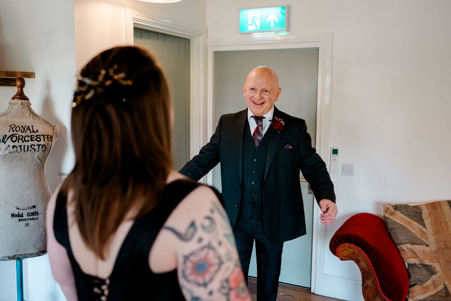 Elderly man in navy suit smiling at tattooed bride during wedding preparations in a bright room.