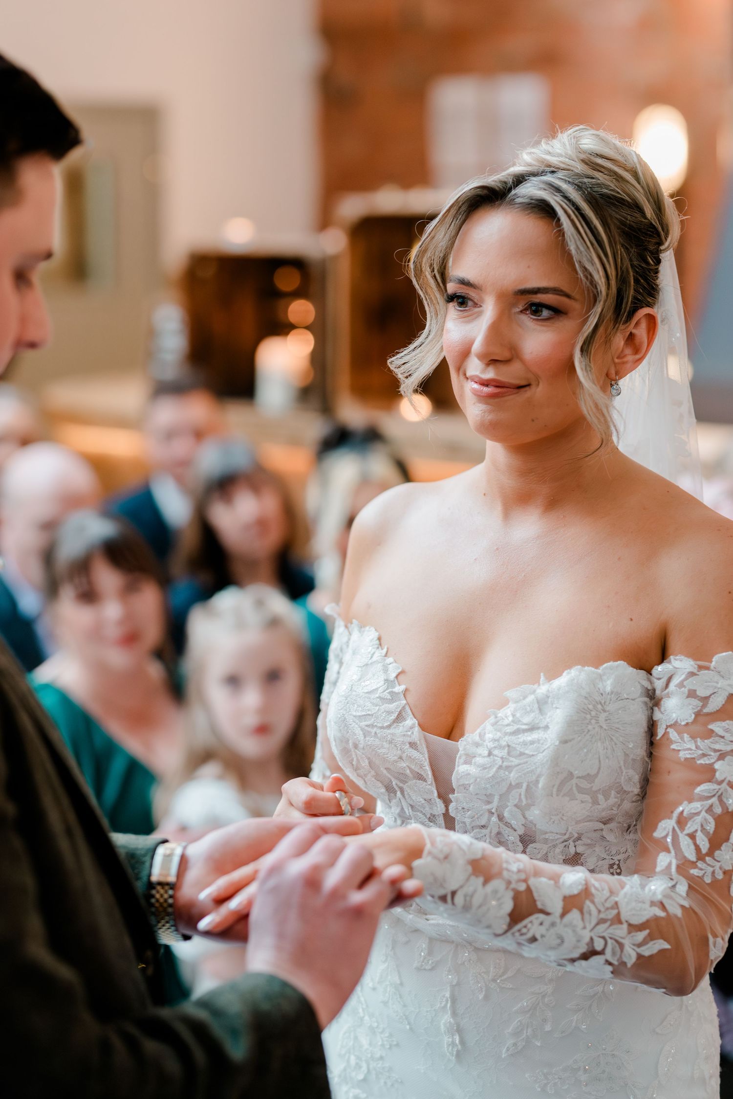 Beautiful bride smiles during ring exchange in stunning off-shoulder lace wedding gown.