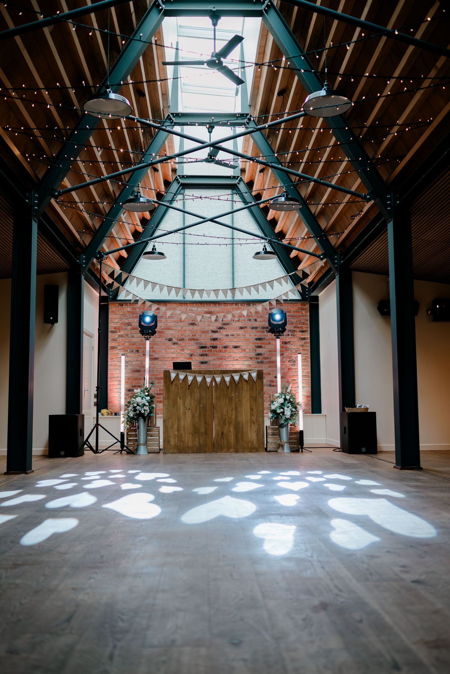 Elegant wedding venue interior with wooden dance floor, rustic DJ booth, brick wall, and decorative bunting lights.