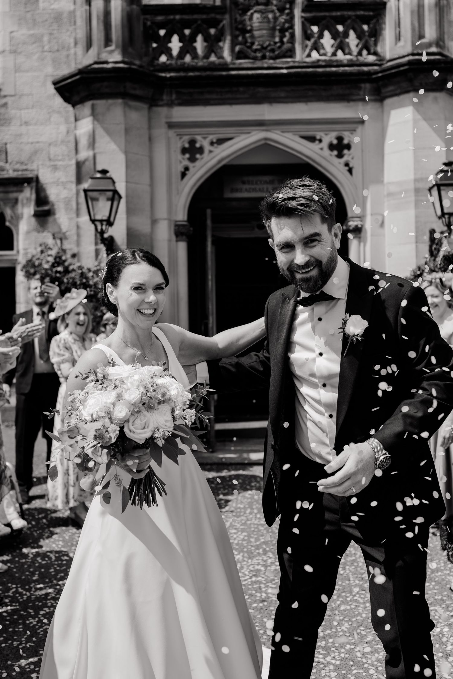 Black and white photo of a smiling bride holding a bouquet and a groom in a tuxedo covered in confetti outside a stone church.