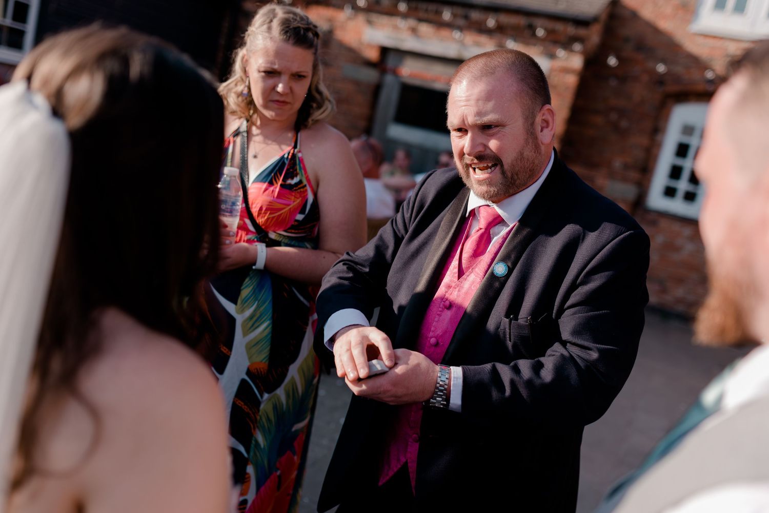 Wedding guest in a pink tie and black suit checks his watch during outdoor reception.