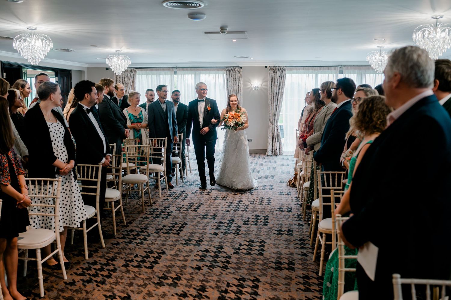 Wide angle view of wedding guests seated in rows at an indoor reception venue with ornate lighting fixtures.