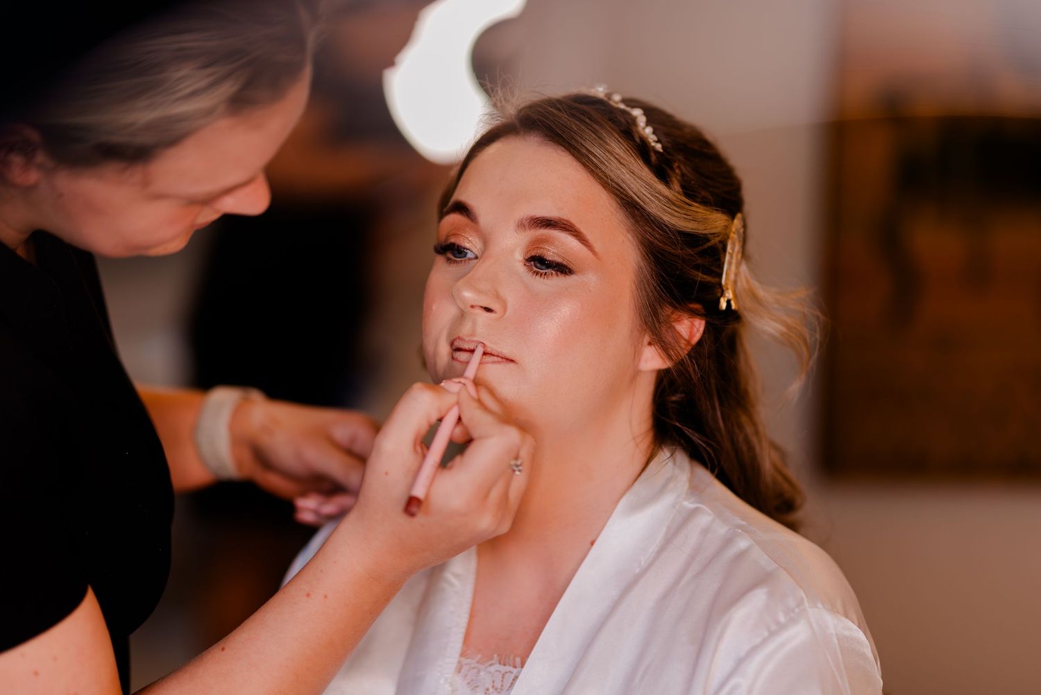 Makeup artist applies lipstick on client wearing white robe during bridal preparation session.