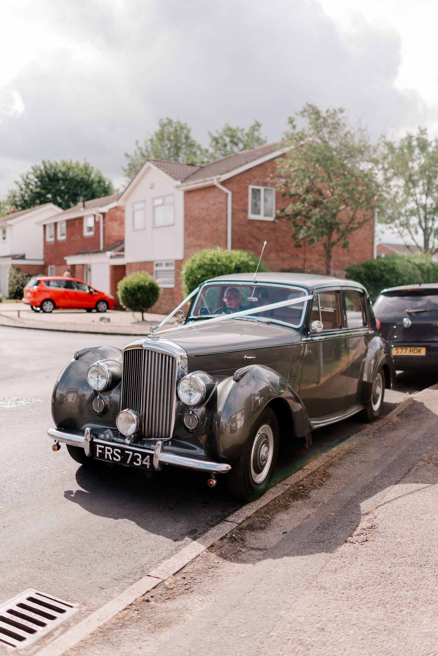 Classic Bentley luxury car parked on residential street with red brick houses and modern vehicles in background.