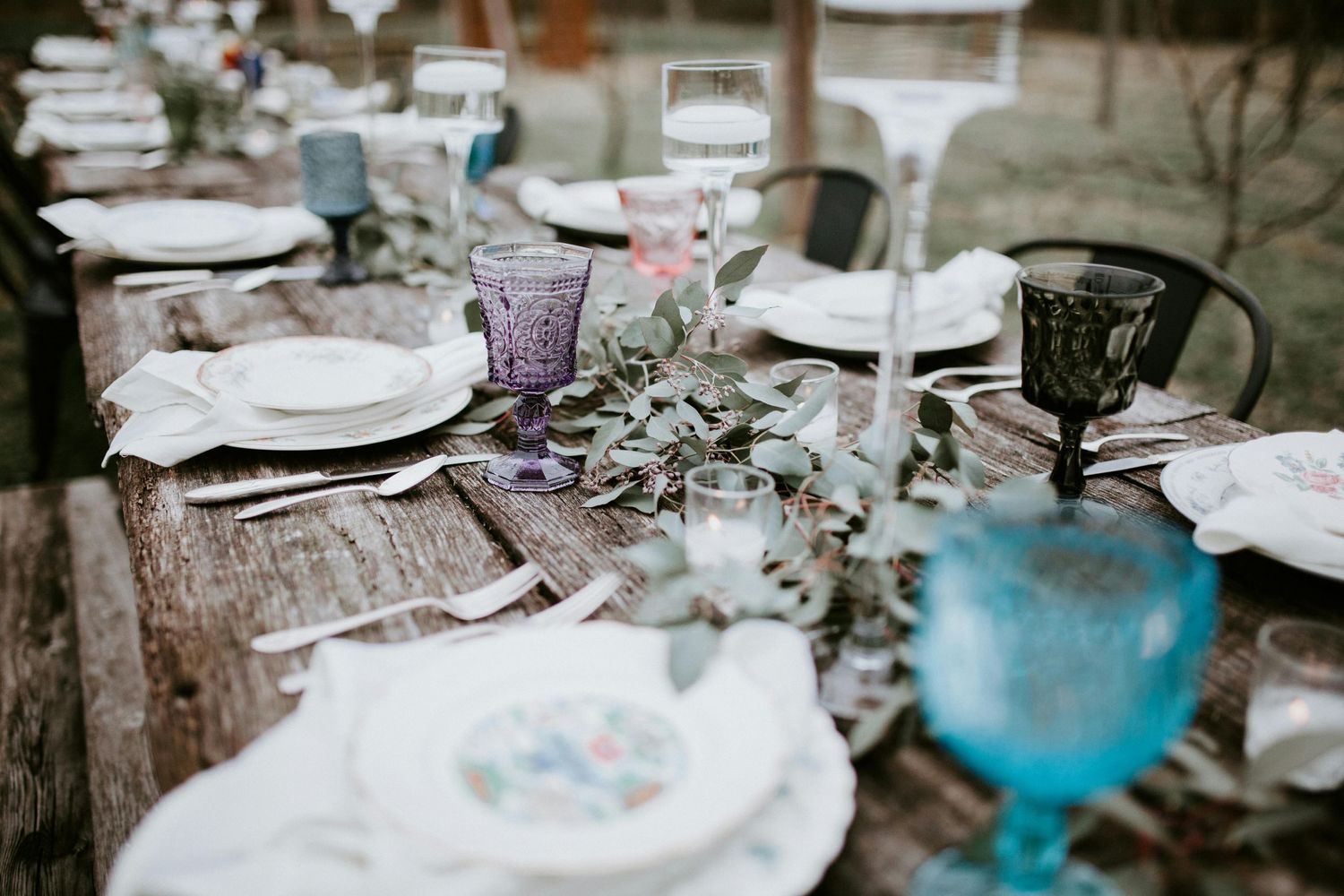 A rustic wedding table setting with white plates, colored glassware, and eucalyptus garland on a wooden table.