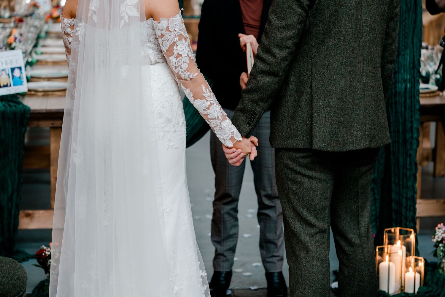Bride and groom hold hands during wedding ceremony with candles and officiant at rustic indoor venue.