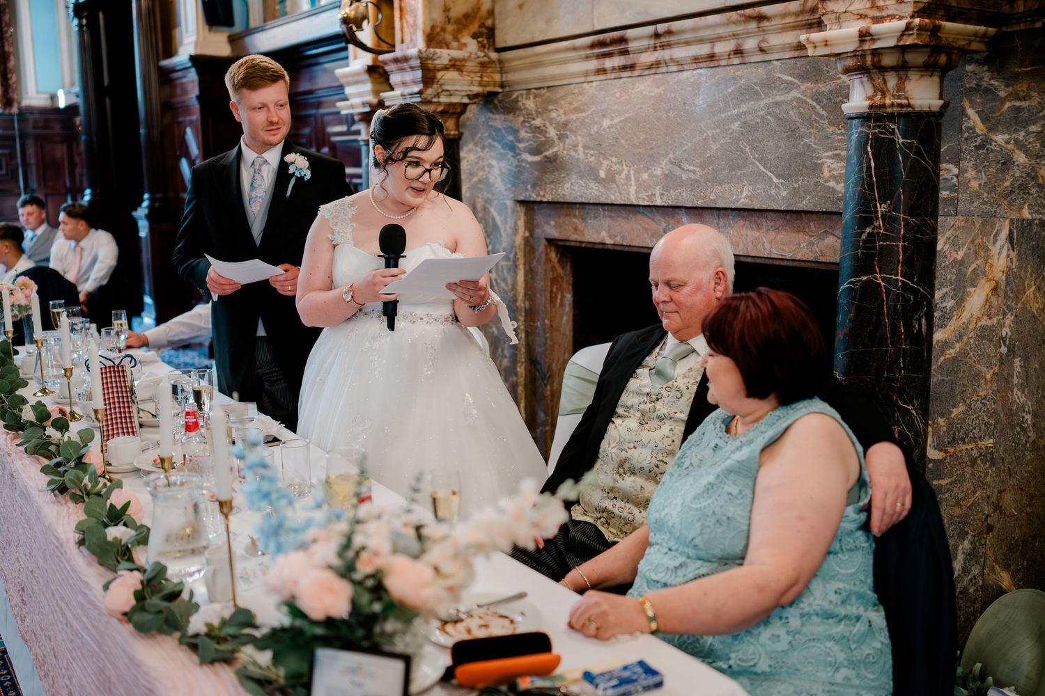 Bride gives speech at wedding reception while groom and seated guests listen near floral centerpieces.