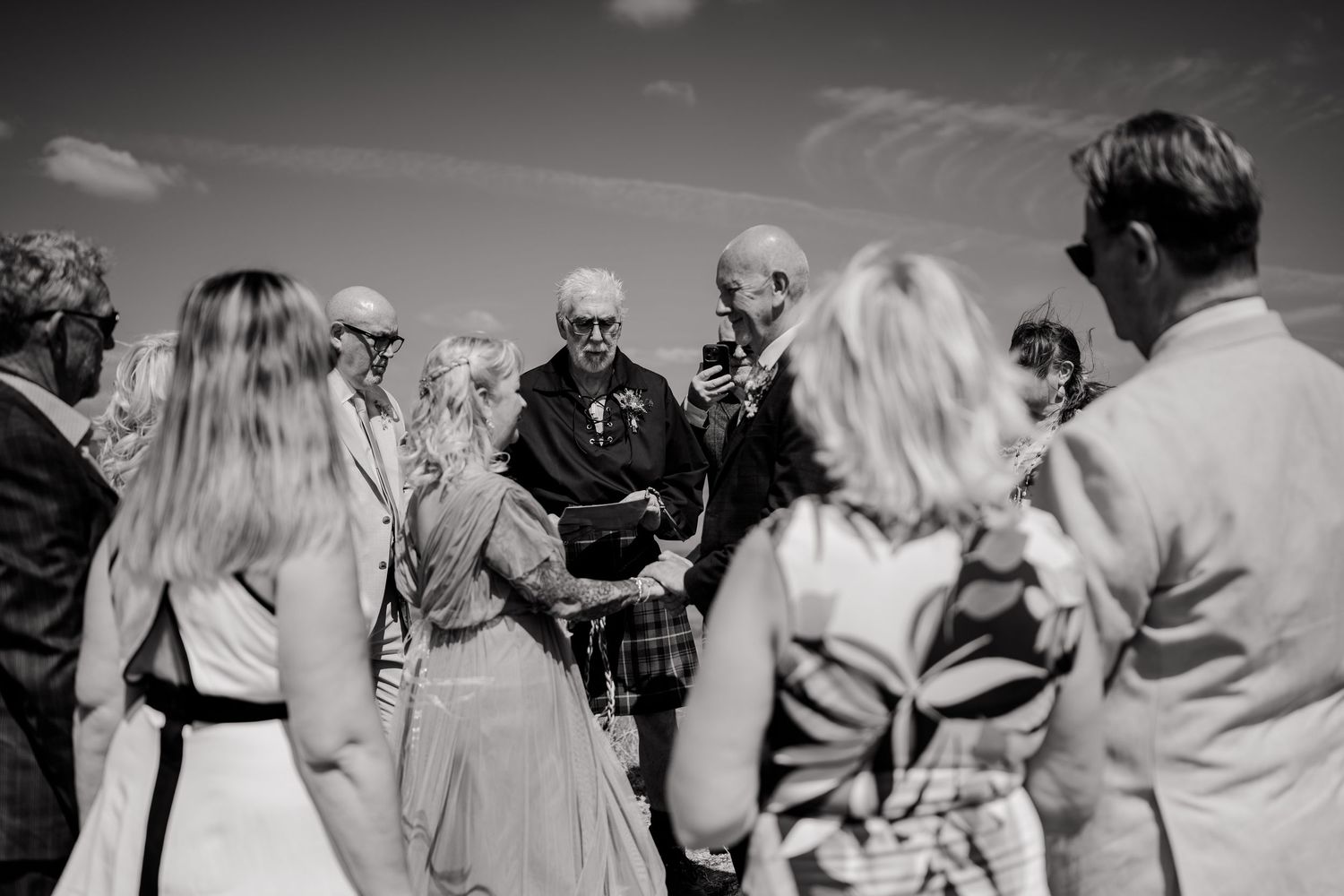 Black and white photo of wedding guests gathered in a circle during an outdoor hilltop ceremony.