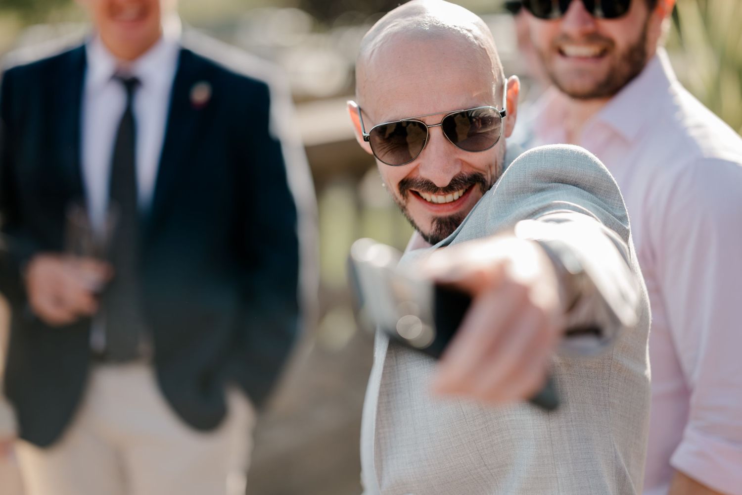 Smiling bald man in sunglasses and a light gray jacket points a camera toward the viewer at an outdoor wedding celebration.