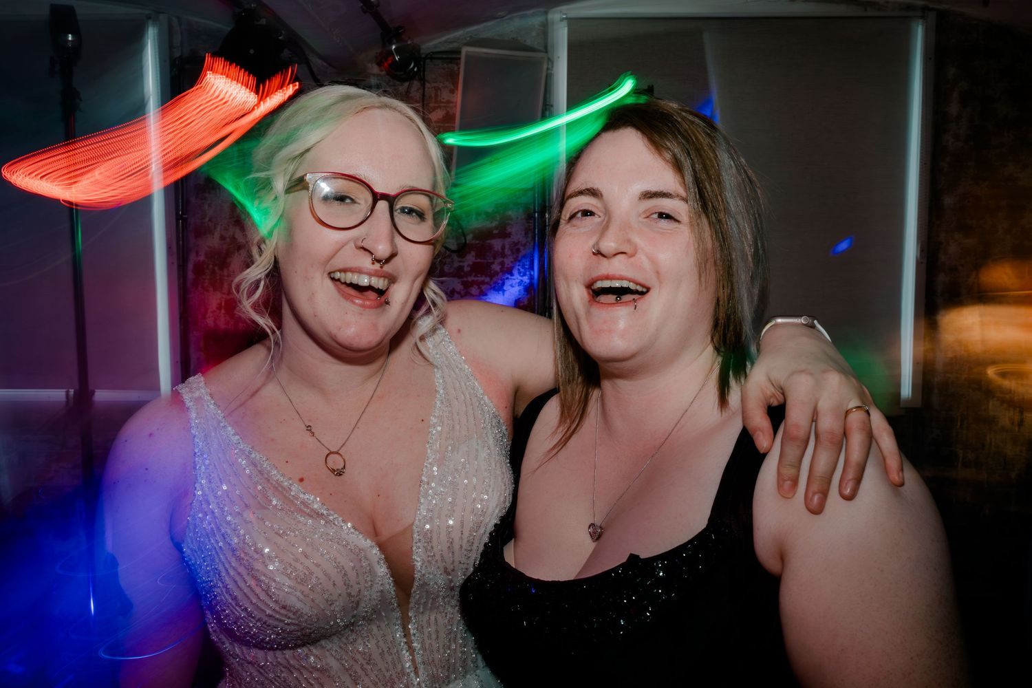 Two smiling women posing together at a neon-lit party venue, one wearing glasses and a sparkly top.