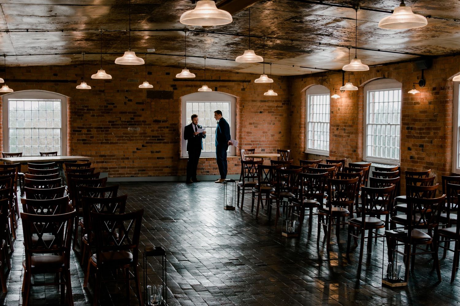 Rustic brick ceremony room with dark wooden chairs, pendant lights, and large windows in a mill venue.