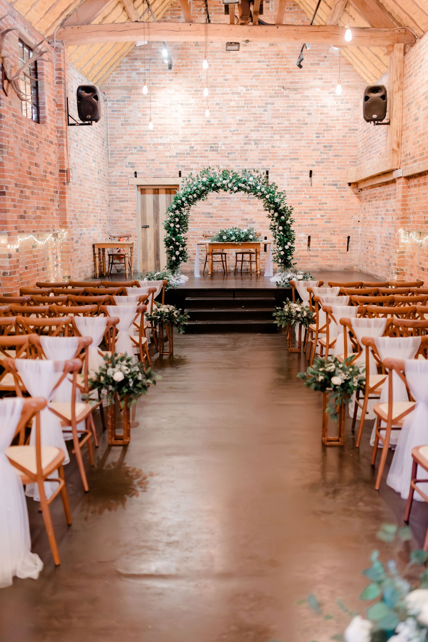 A rustic wedding ceremony setup with a floral arch and wooden chairs in a brick-walled venue.
