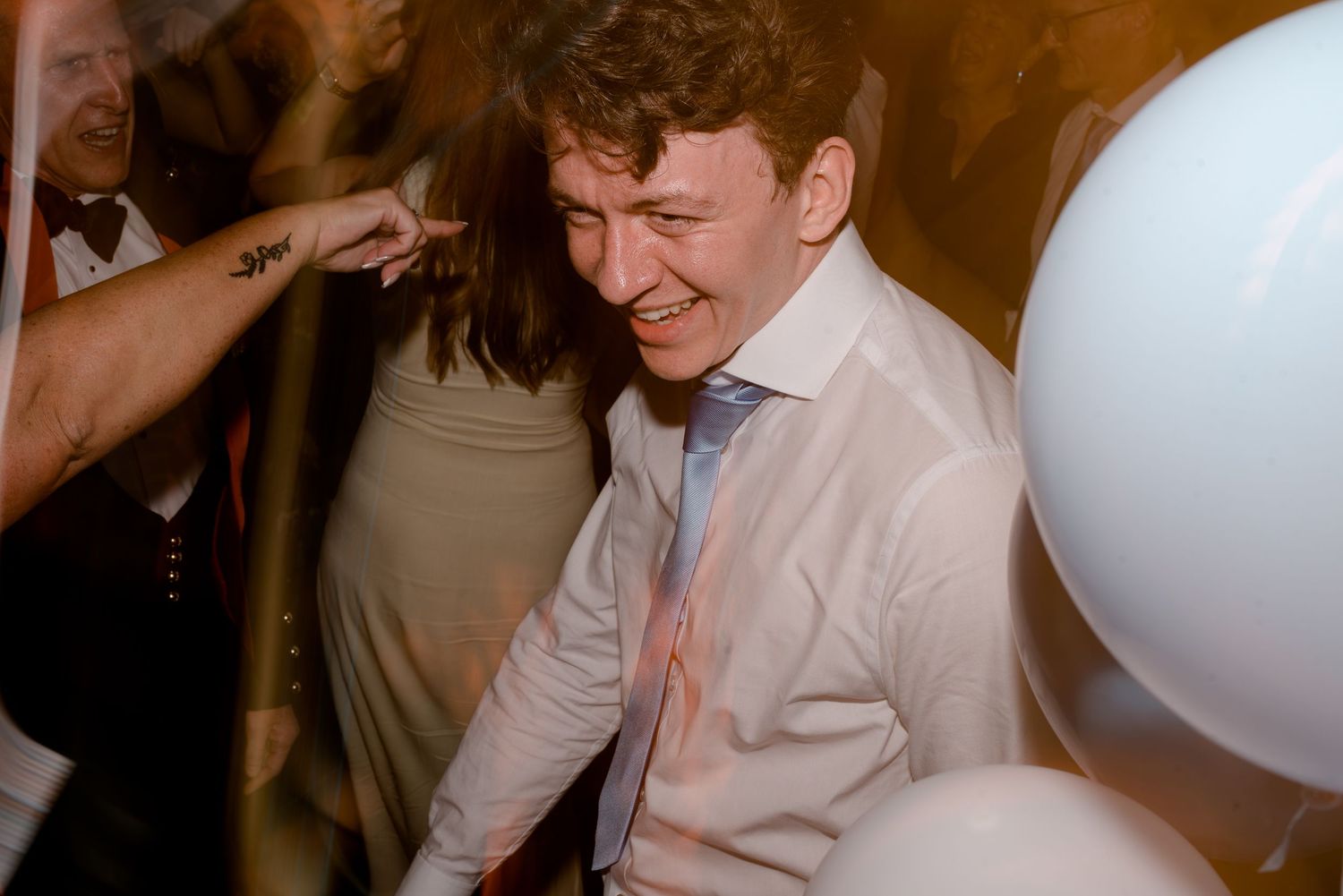 A person in a white dress shirt laughs joyfully while dancing at a celebration with a balloon visible in the background.