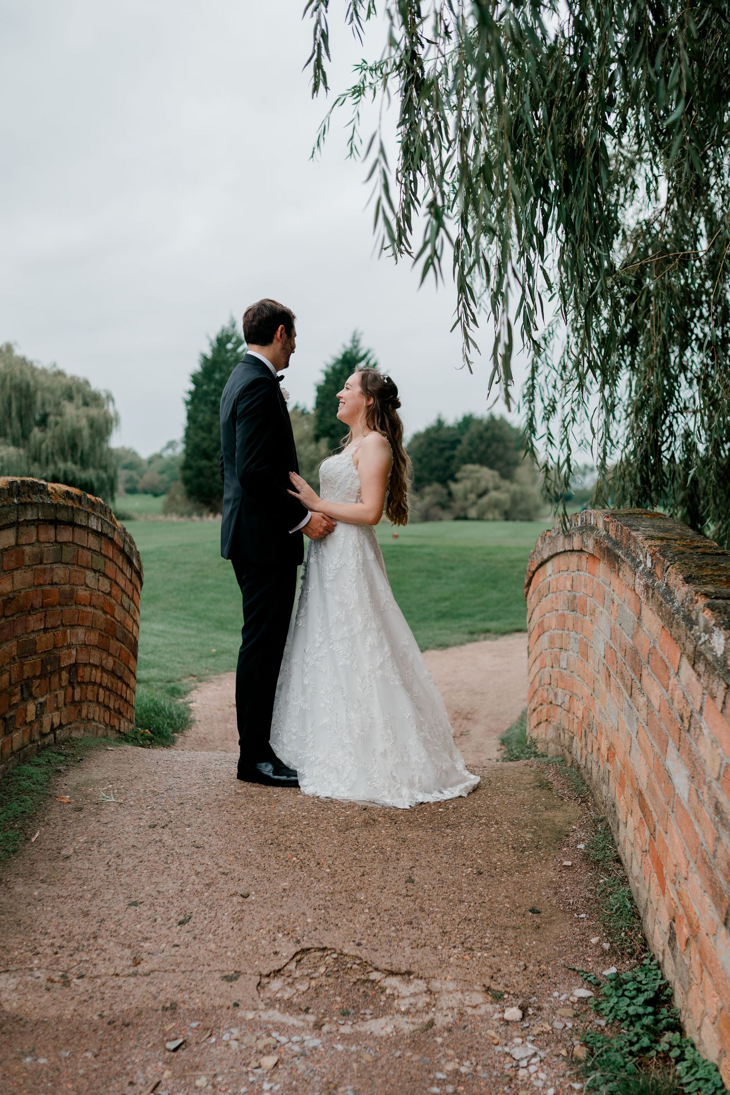 A bride and groom share an intimate moment under a weeping willow tree on a stone path with brick walls.