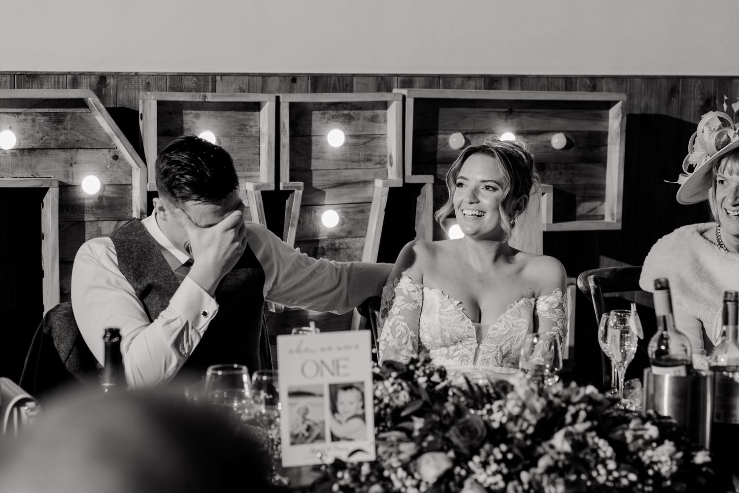 Black and white photo of groom covering face while bride smiles during wedding speeches.