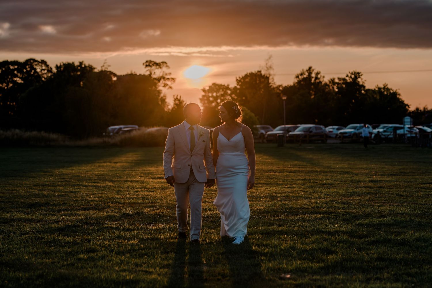 Couple walking hand in hand through a field at sunset with cars parked in background.
