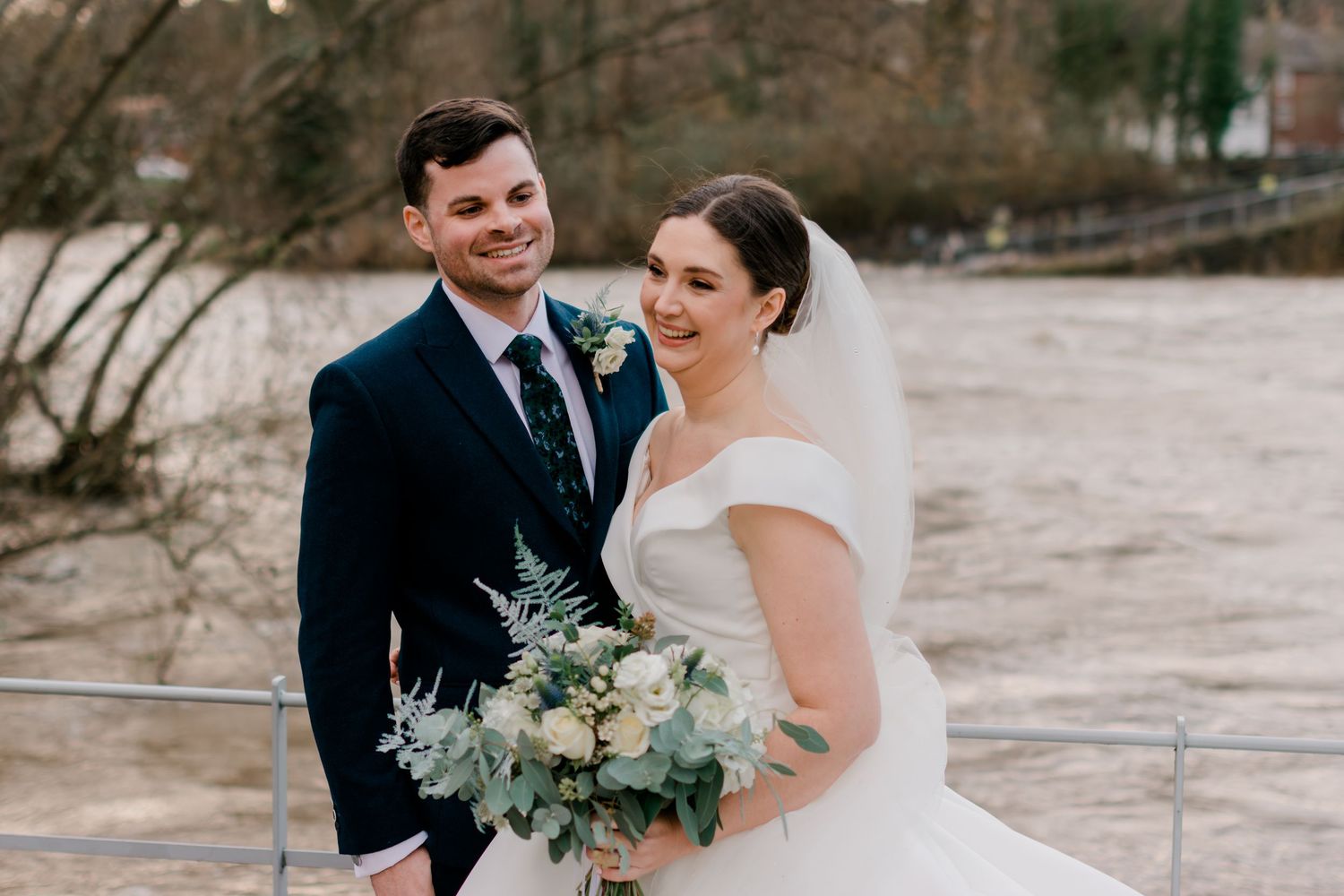 Smiling bride and groom posing together by a river holding a white and green bouquet.