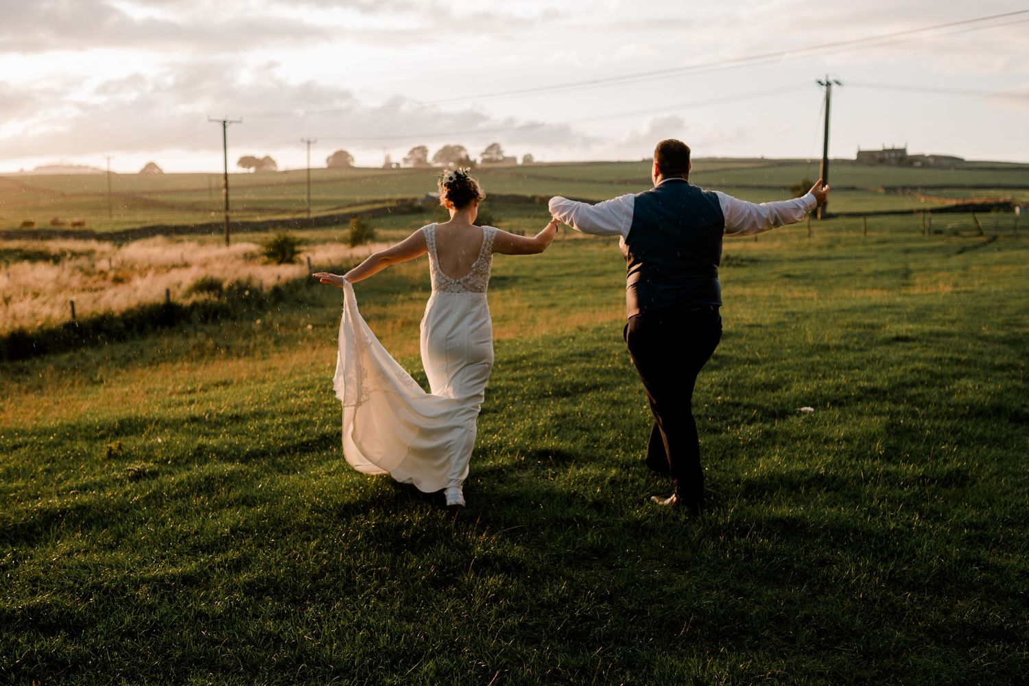 Bride and groom walking hand in hand across a green field at sunset, wedding dress flowing in the breeze.